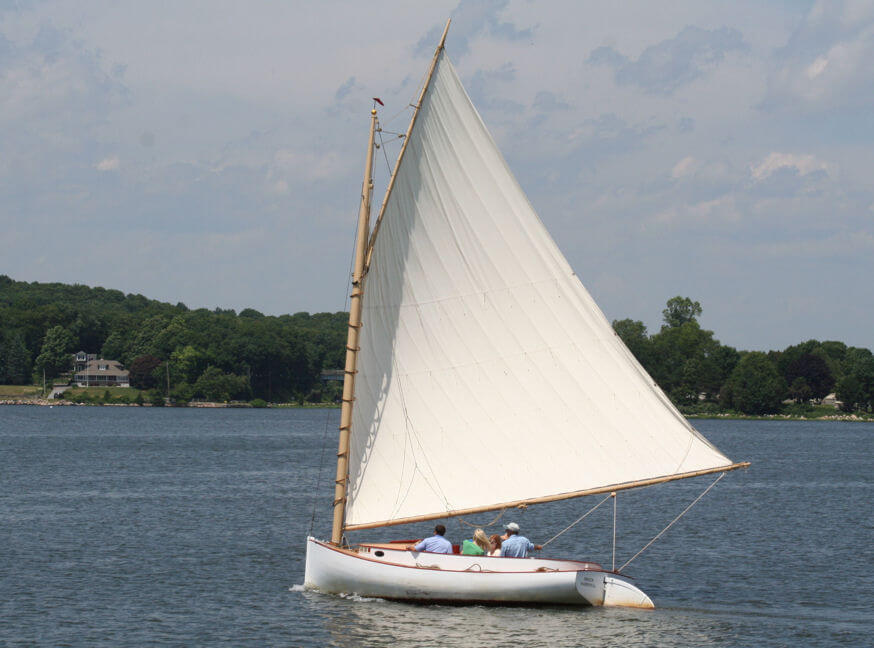 White cat boat with gaff rig on lake and three people in it