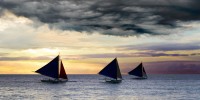 3 Sailboats with dark sails under dark clouds at sunset