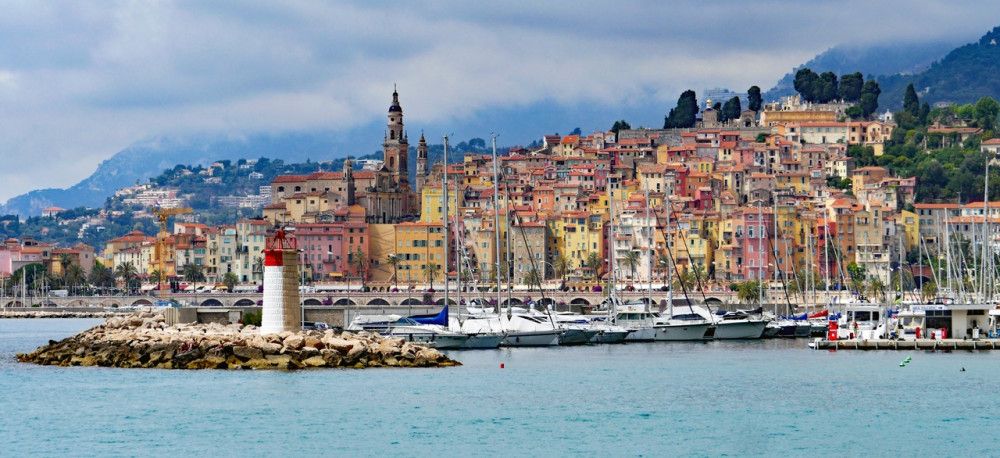 Coastline filled with colorful houses, with a small lighthouse and sailing yachts offshore and a mountain in the back