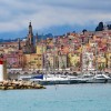 Coastline filled with colorful houses, with a small lighthouse and sailing yachts offshore and a mountain in the back