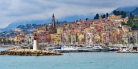 Coastline filled with colorful houses, with a small lighthouse and sailing yachts offshore and a mountain in the back